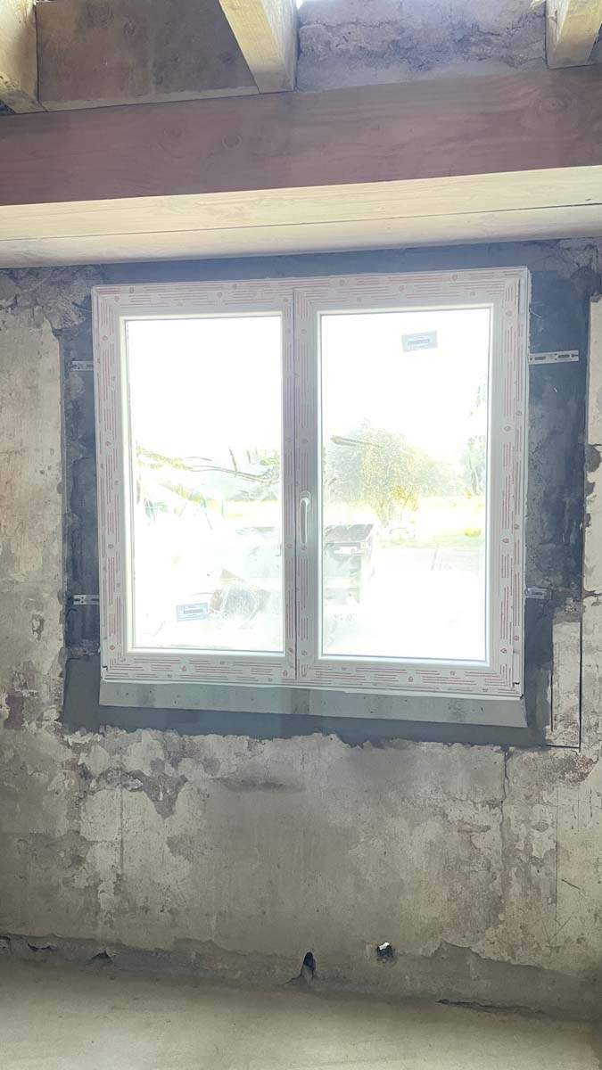 Fenêtre en PVC blanc à double vitrage et à deux battants, installée dans une ouverture de mur en béton brut Josselin